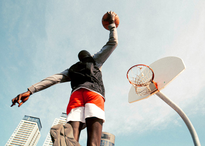 Man in black shirt and red and white shorts jumping up to dunk a basketball on a rim with skyscrapers in the backdrop.